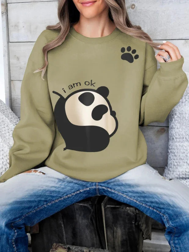 Woman wearing olive green sweatshirt with cute panda graphic and 'I am ok' text, paired with blue jeans