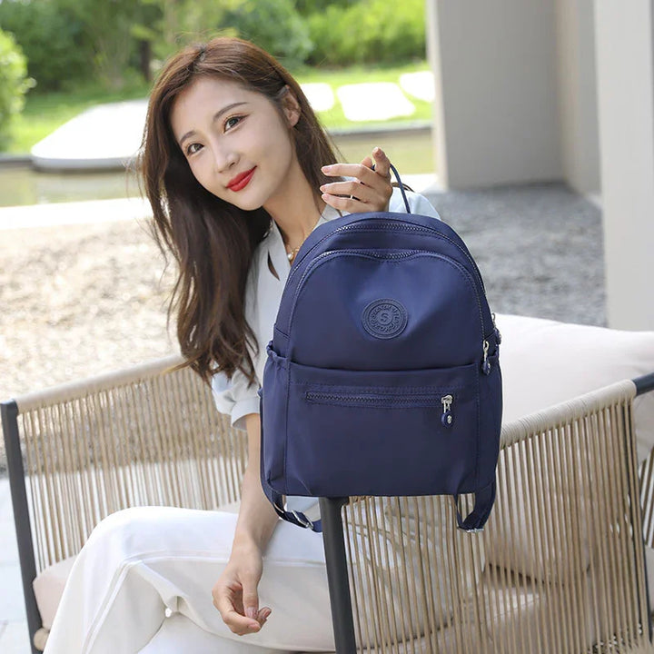 Smiling woman in white outfit sitting outdoors holding a navy blue casual backpack on chair