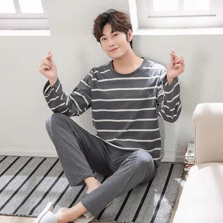 Young man sitting on striped rug wearing grey striped long-sleeve shirt and grey pants in bright room