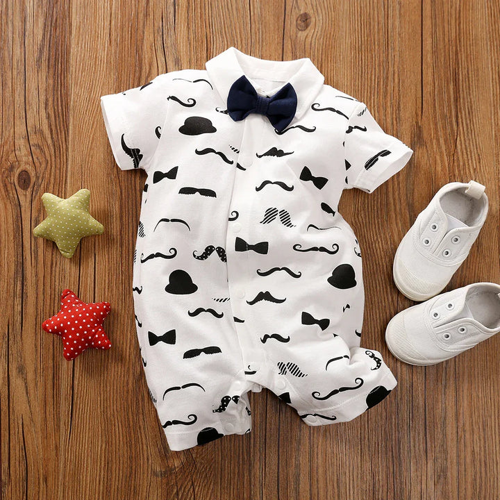 Baby boy romper with black bow tie and mustache pattern, white shoes, and colorful star toys on wooden floor
