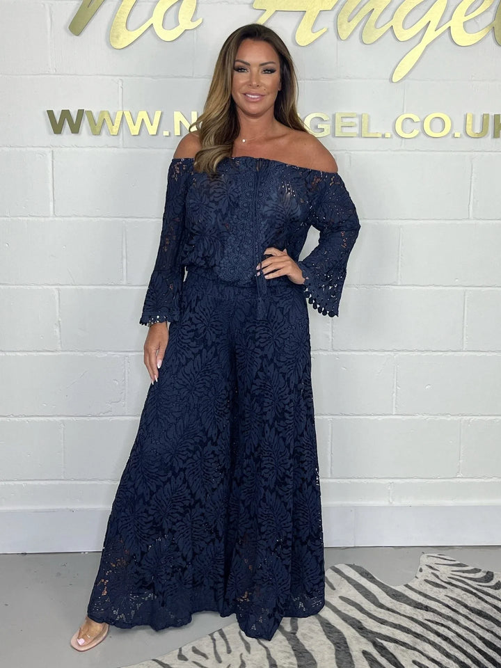 Woman wearing navy blue off-shoulder lace jumpsuit with flared pants standing against white brick wall