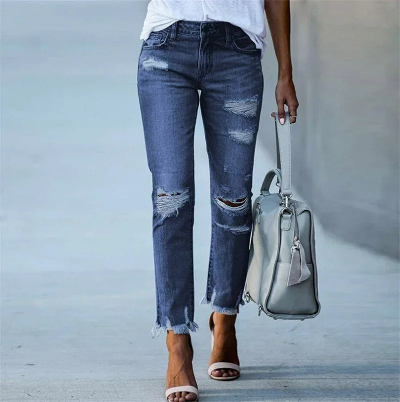 Woman wearing distressed blue jeans and white sandals carrying a light gray handbag on a neutral background