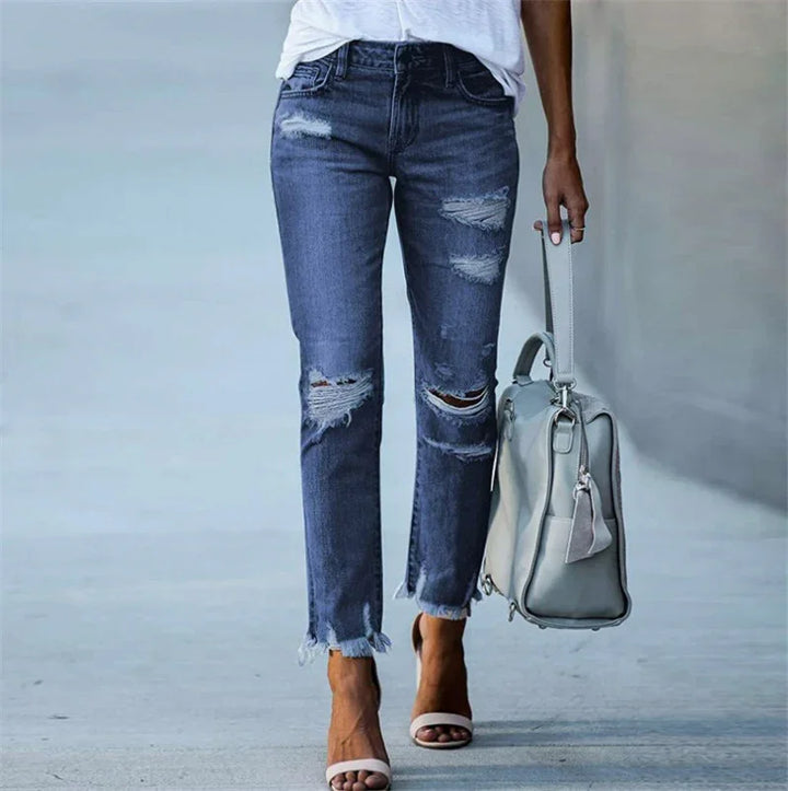 Woman wearing distressed blue jeans and white sandals carrying a light gray handbag on a neutral background