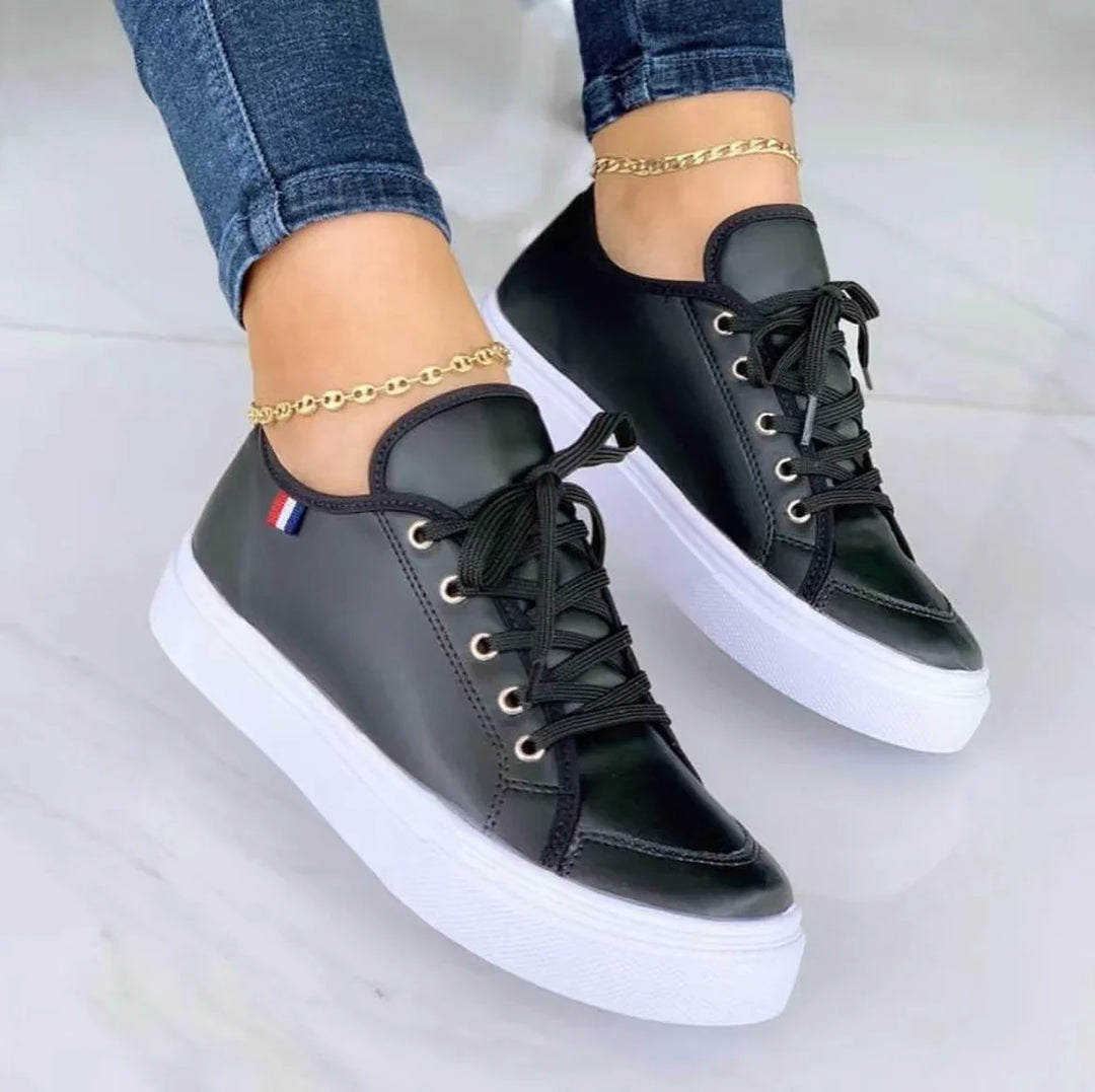 Black leather low-top sneakers with white thick soles worn with gold anklets and blue jeans on white floor