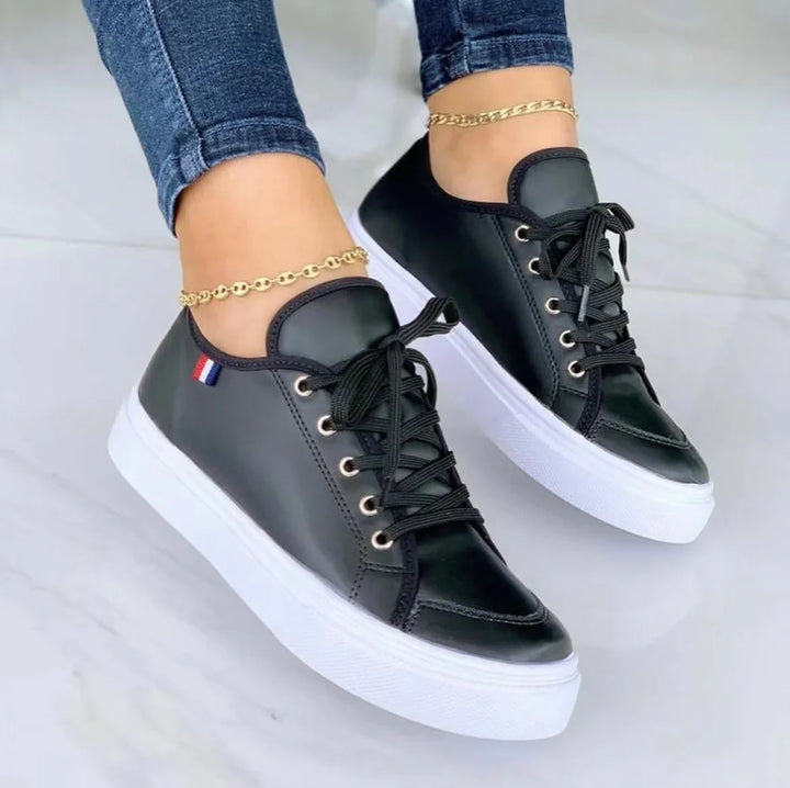Black leather low-top sneakers with white thick soles worn with gold anklets and blue jeans on white floor