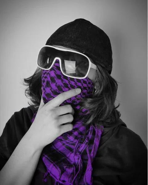 Person wearing black cap and large white sunglasses with purple checkered scarf covering lower face