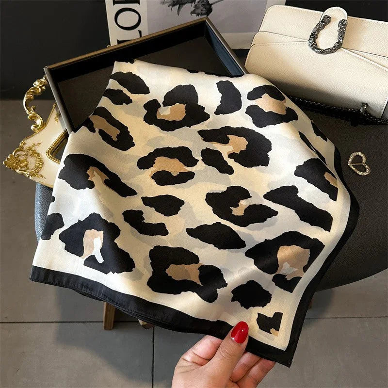 Hand holding leopard print silk scarf with black border on table next to white handbag and jewelry
