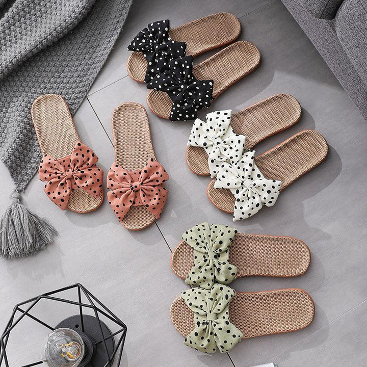 Four pairs of woven slippers with large polka dot bows in black, white, pink, and green on a gray tile floor