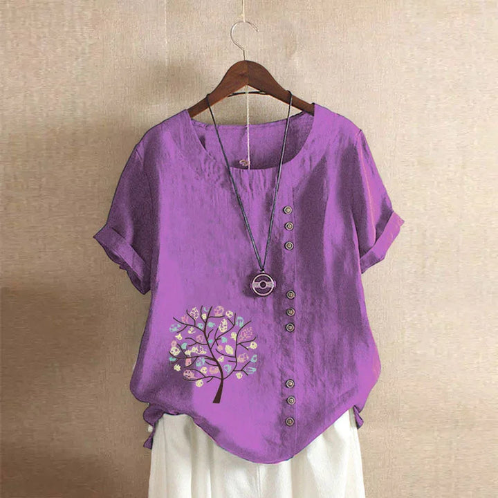 Purple short sleeve blouse with front buttons and tree print paired with white pants on wooden hanger