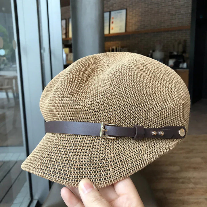 Hand holding beige woven newsboy cap with brown leather strap in modern cafe setting