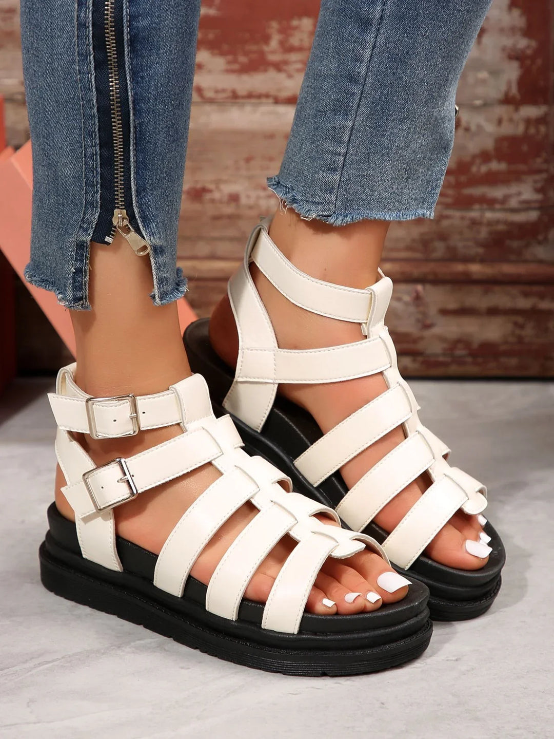 White strappy leather gladiator sandals with buckles on feet wearing blue denim jeans with zipper detail