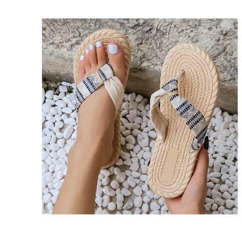 beige braided sole flip flops with black and white patterned straps worn on feet with white pedicure on white pebbles