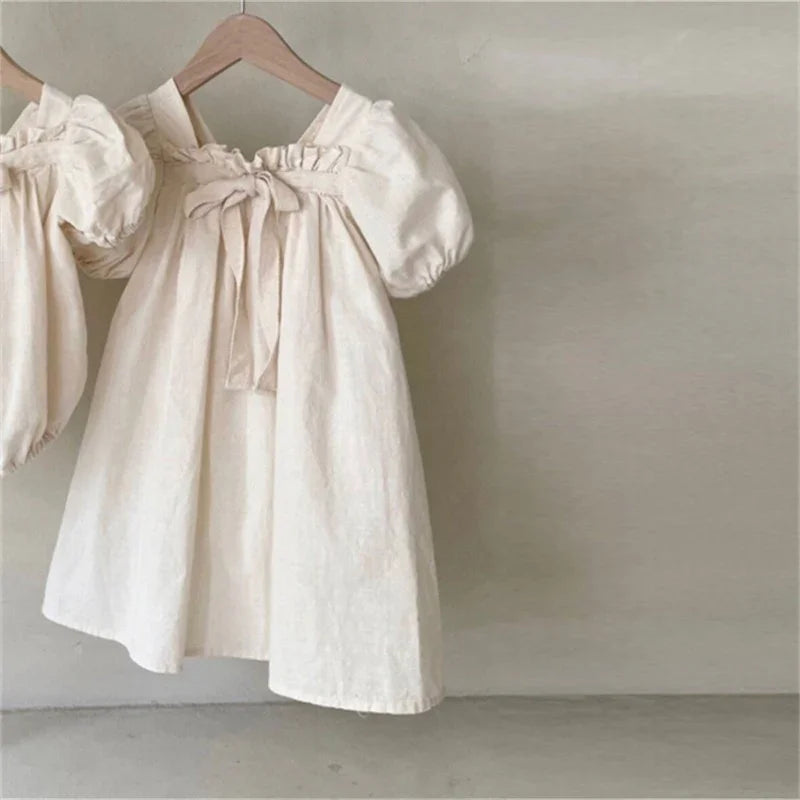 Cream-colored baby dress with puff sleeves and front bow detail on wooden hanger