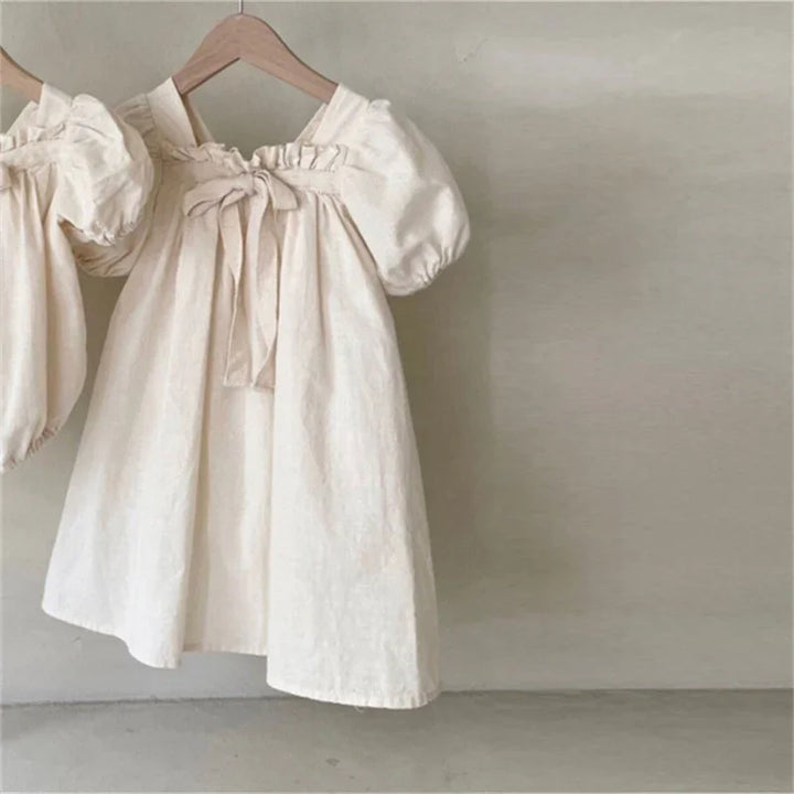 Cream-colored baby dress with puff sleeves and front bow detail on wooden hanger