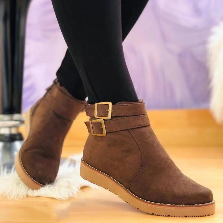 Brown suede ankle boots with double gold buckle straps worn with black leggings on wooden floor