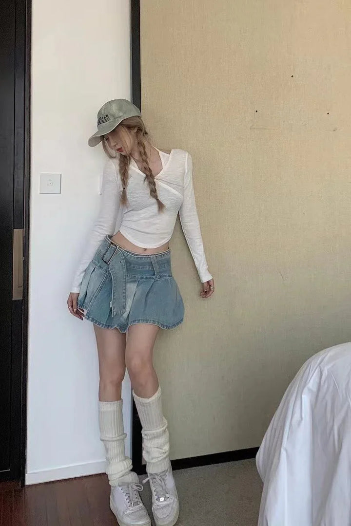 Young woman with braided hair wearing a white long-sleeve top, denim skirt, slouchy socks, white sneakers, and a gray cap indoors