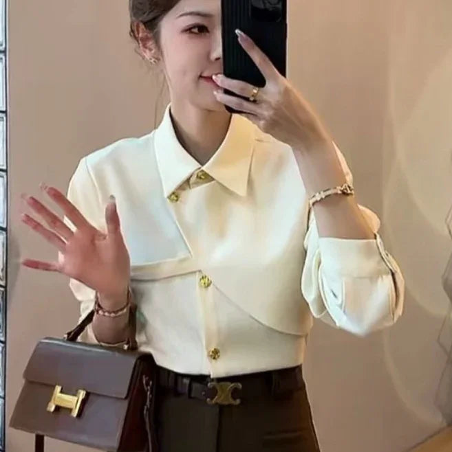 Woman taking mirror selfie wearing cream asymmetrical buttoned blouse, black pants, and holding brown leather handbag