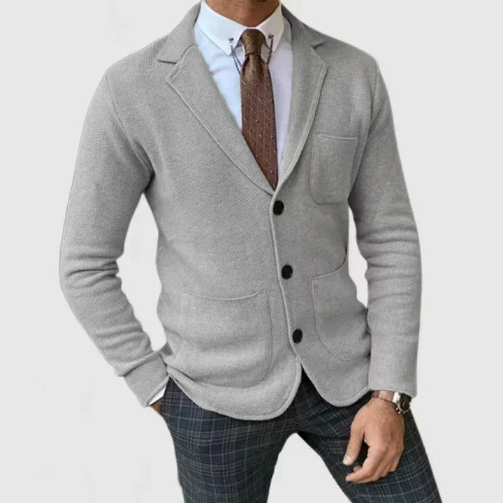 Man wearing gray knit blazer with black buttons, white shirt, brown tie, and plaid pants