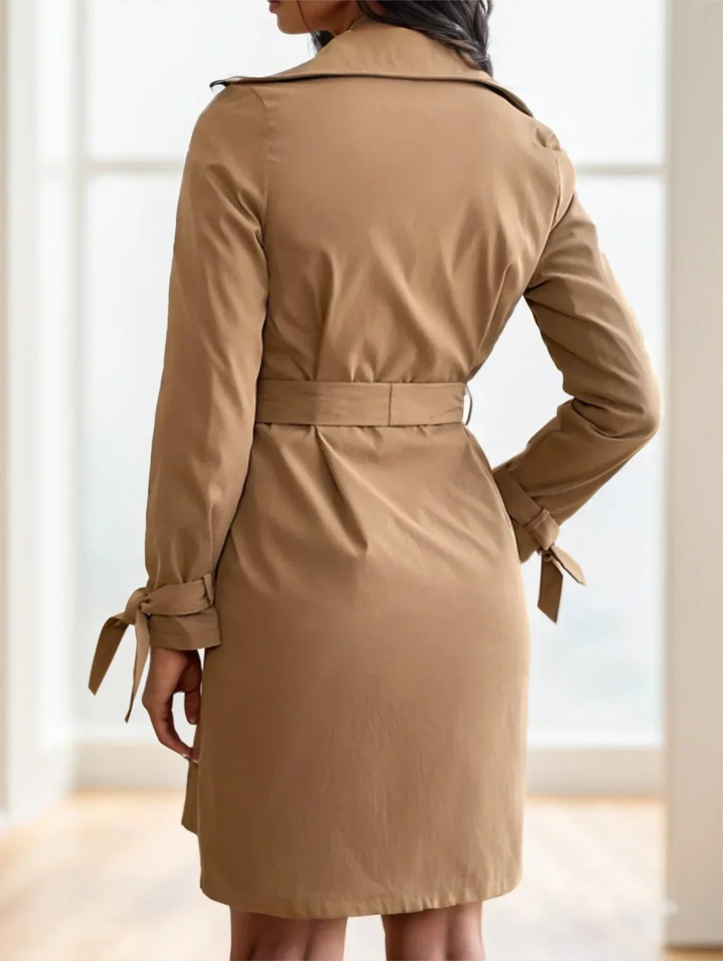 Back view of woman wearing camel brown belted trench coat with bow-tied cuffs indoors