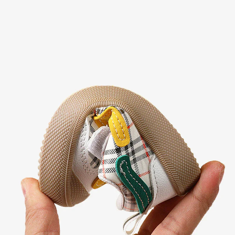 Flexible white baby sneaker with plaid pattern and yellow and green accents held in hand
