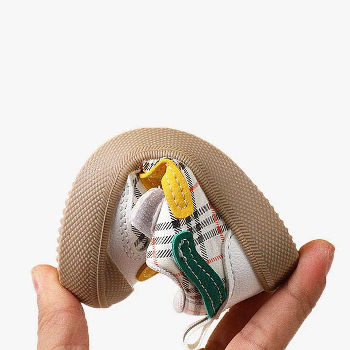 Flexible white baby sneaker with plaid pattern and yellow and green accents held in hand