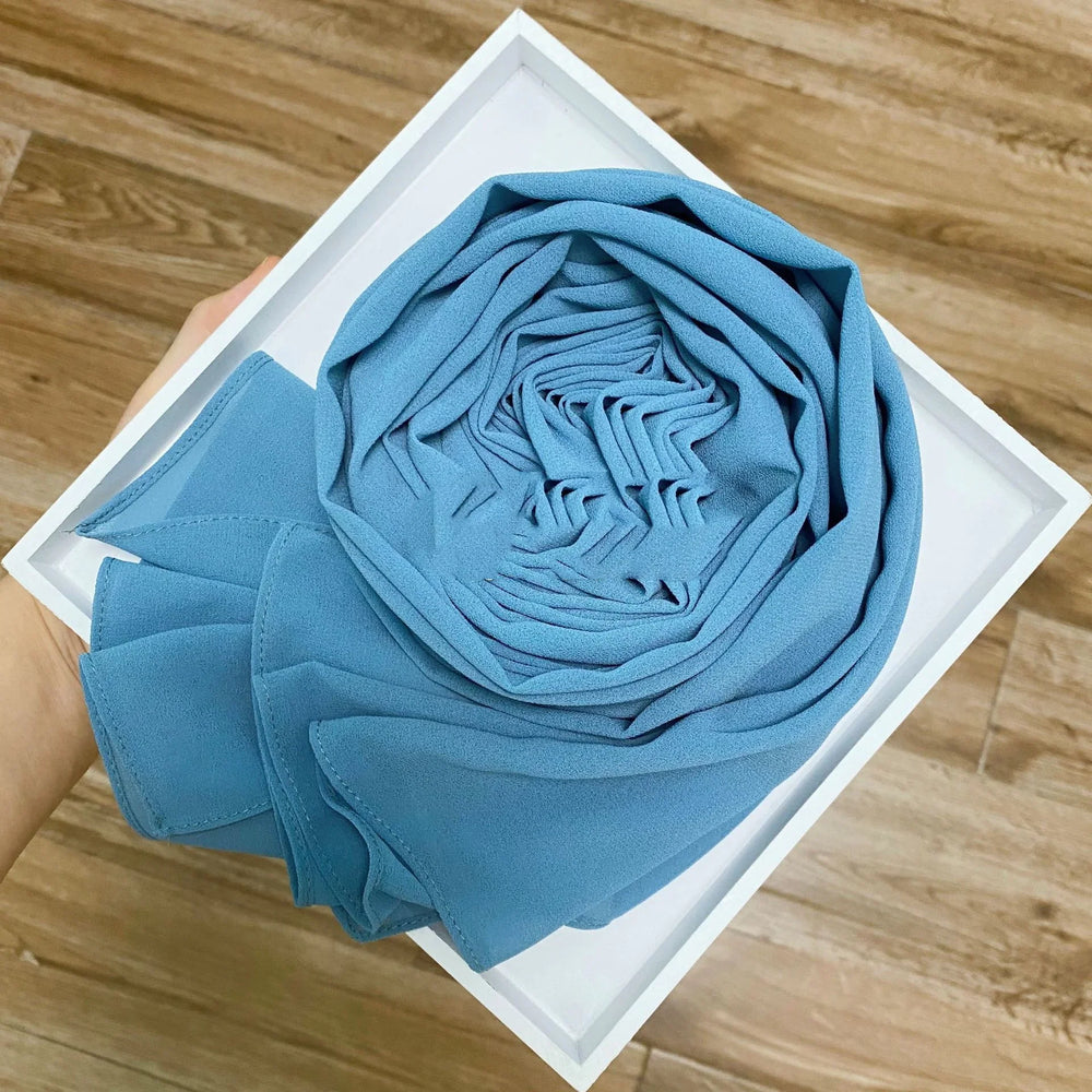 Hand holding white box with neatly rolled light blue chiffon fabric scarf on wooden floor