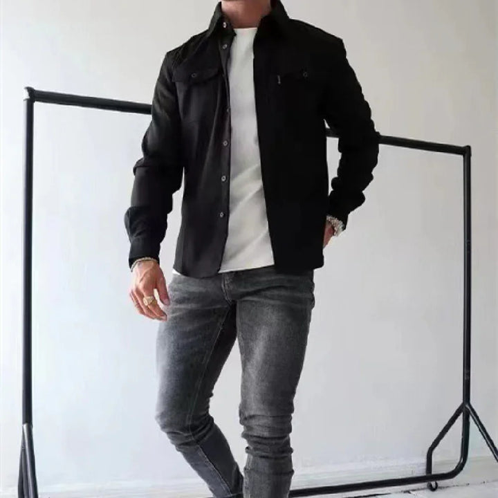 Man wearing black jacket, white shirt, and gray jeans standing indoors by clothing rack