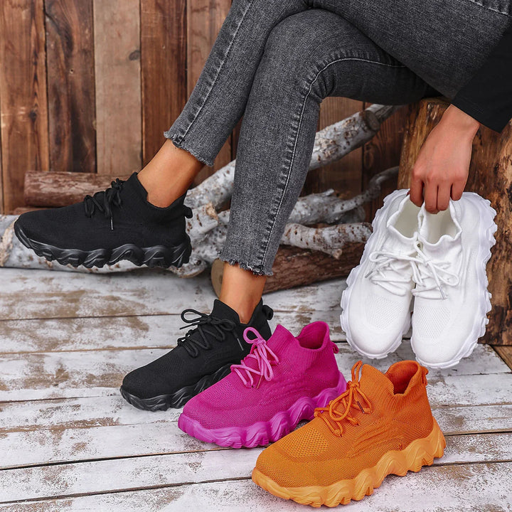 Person wearing black knitted sneakers and holding white sneakers with pink and orange pairs on wooden floor