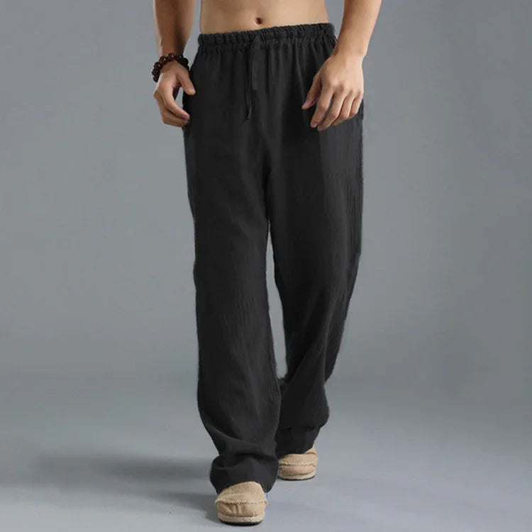 Man wearing loose black linen pants with elastic waistband and beige slippers on gray background