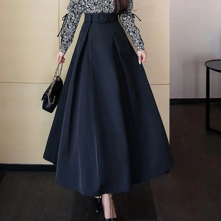 Woman wearing high-waisted black A-line skirt with belt, floral long-sleeve top, and black handbag