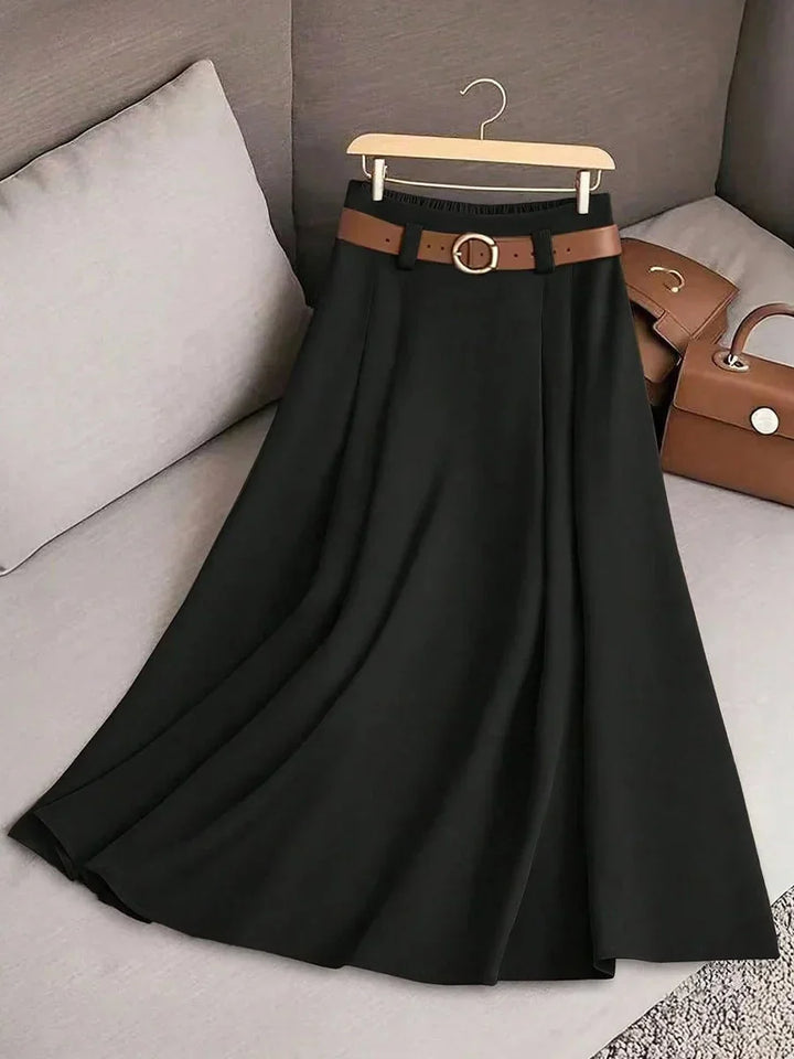 Black midi skirt with pleats and brown belt displayed on wooden hanger on gray sofa with brown handbags
