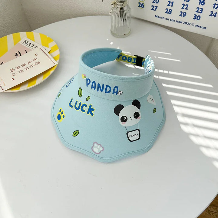 Light blue panda-themed sun visor with 'PANDA' and 'Luck' text on white table with striped yellow plate and calendar