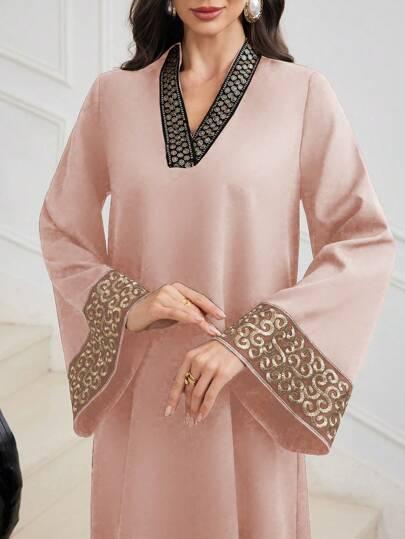 Woman wearing light pink abaya with gold and black embroidered cuffs and neckline in elegant indoor setting