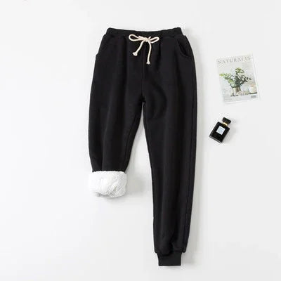 Black fleece-lined jogger pants with white drawstring, displayed with a small plant magazine and perfume bottle on white background