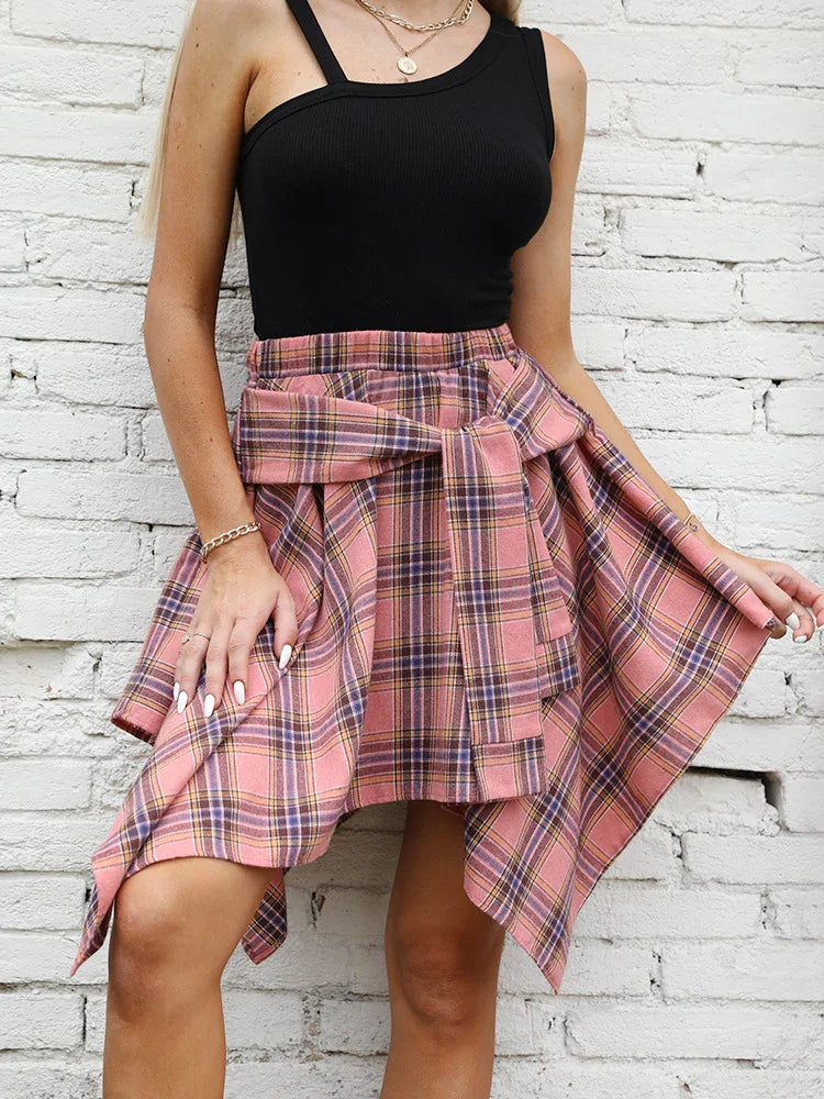 Woman wearing a black tank top and pink plaid handkerchief skirt with tied waist detail against white brick wall