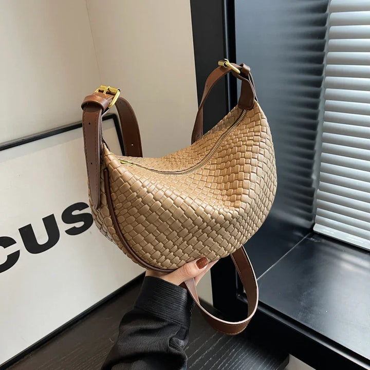 Hand holding a tan woven leather shoulder bag with brown adjustable straps near a window with blinds
