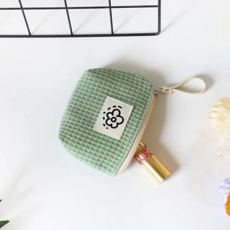 Small green waffle-textured cosmetic pouch with flower logo holding gold lipstick on white surface