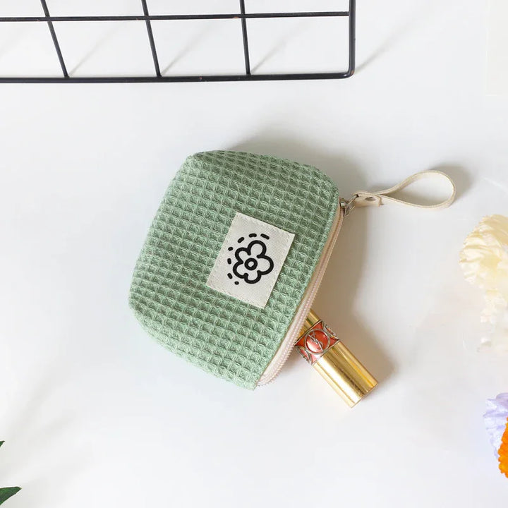 Small green waffle-textured cosmetic pouch with flower logo holding gold lipstick on white surface