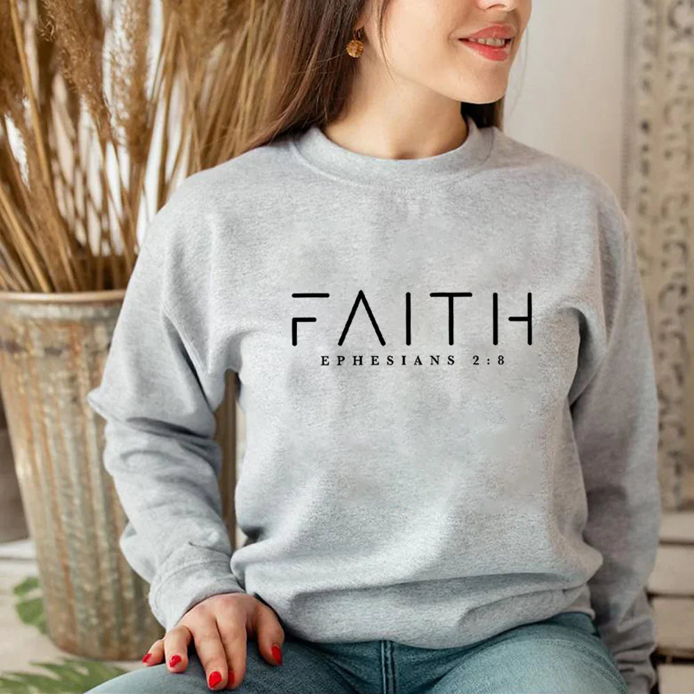 Woman wearing light gray sweatshirt with FAITH and Ephesians 2:8 text, sitting indoors with dried plants