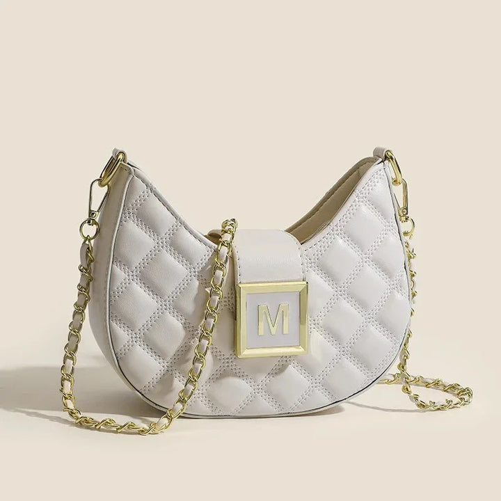 White quilted leather handbag with gold chain strap and gold M logo clasp on beige background