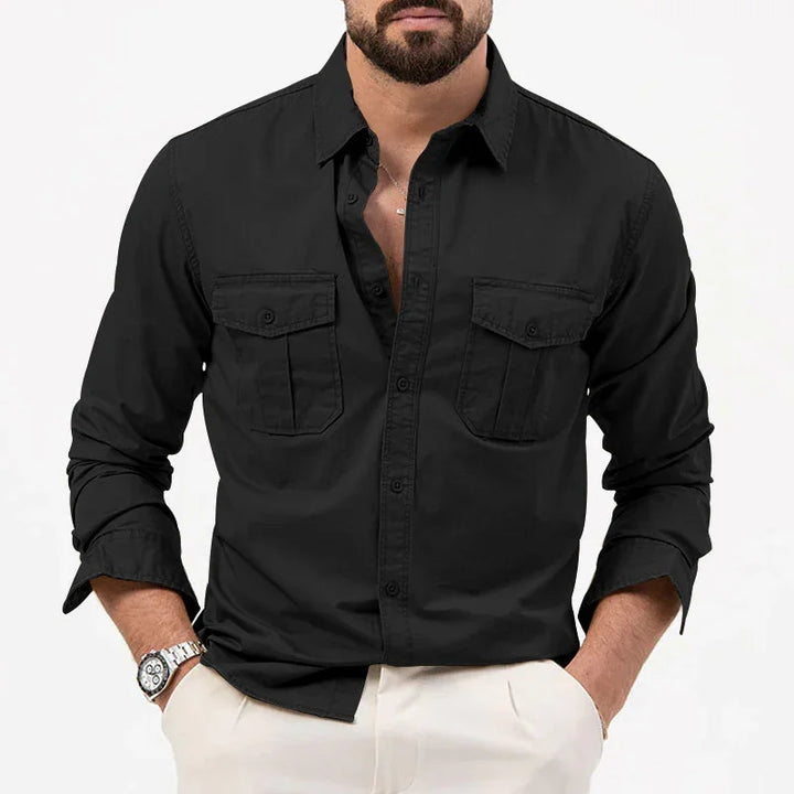 Man wearing black button-up shirt with two chest pockets and white pants, accessorized with watch and necklace