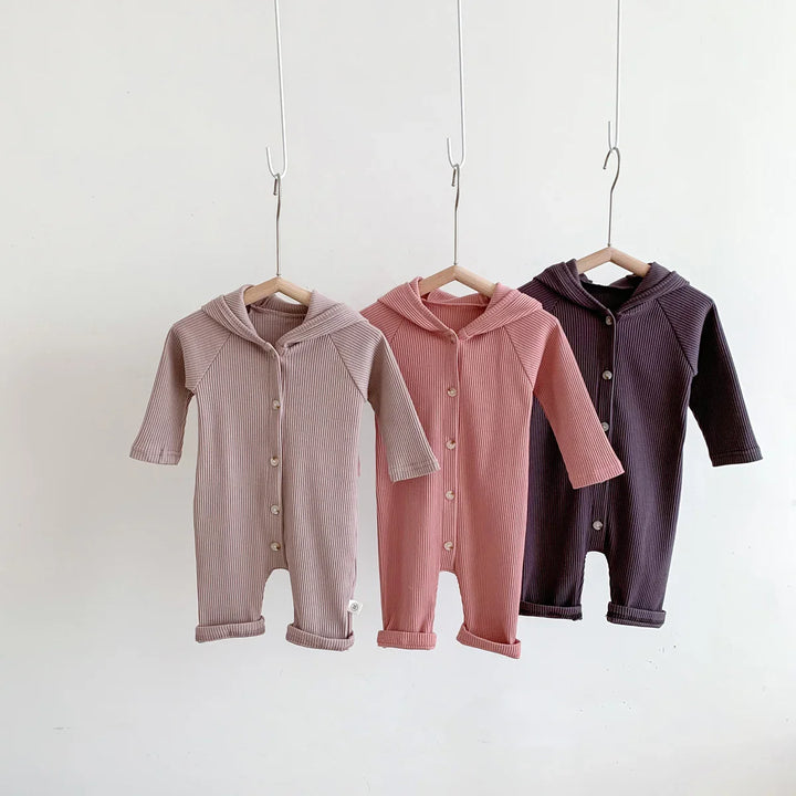 Three ribbed cotton baby hooded onesies in beige, pink, and dark purple hanging on wooden hangers