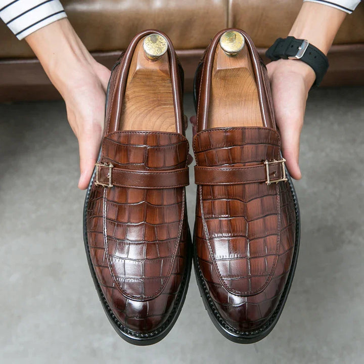 Pair of brown crocodile leather dress loafers with buckles, held by hands over gray floor