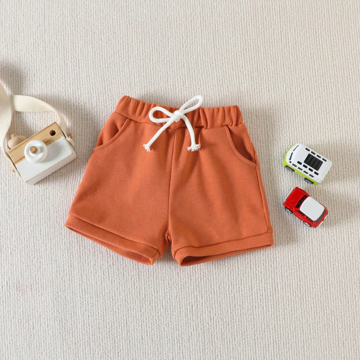 Orange toddler shorts with white drawstring on beige carpet with wooden toy camera and two toy cars
