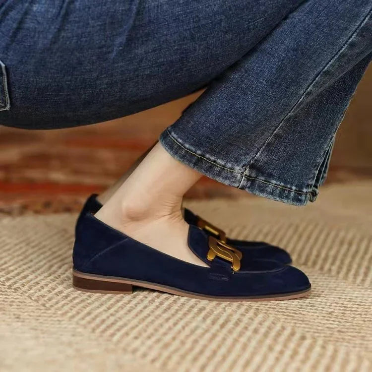 Close-up of navy blue suede loafers with gold buckle detail worn with blue jeans on a textured rug