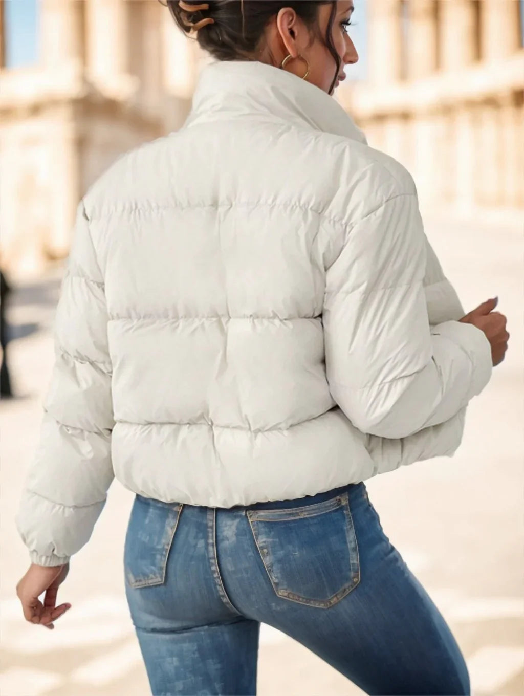 Woman wearing a white puffer jacket and blue jeans outdoors in a sunny urban setting