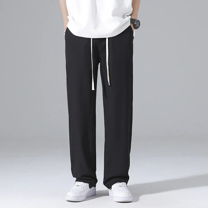 Man wearing black loose fit pants with white drawstrings and white sneakers against gray background