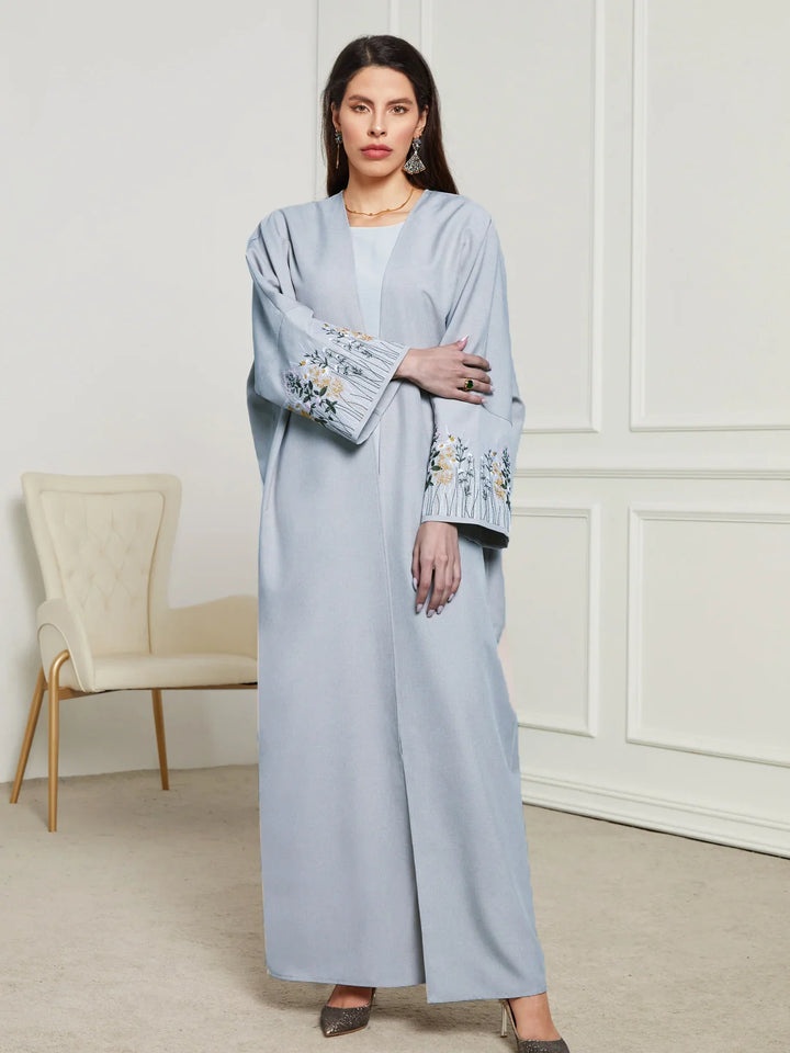 Woman in light blue long abaya with floral embroidery on sleeves, standing in modern room with white walls and beige chair