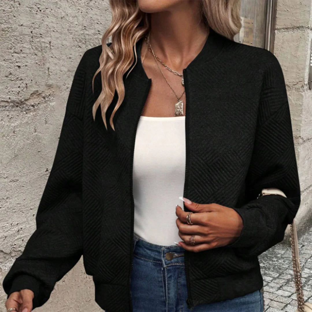 Woman wearing black textured bomber jacket over white top with blue jeans, urban wall background