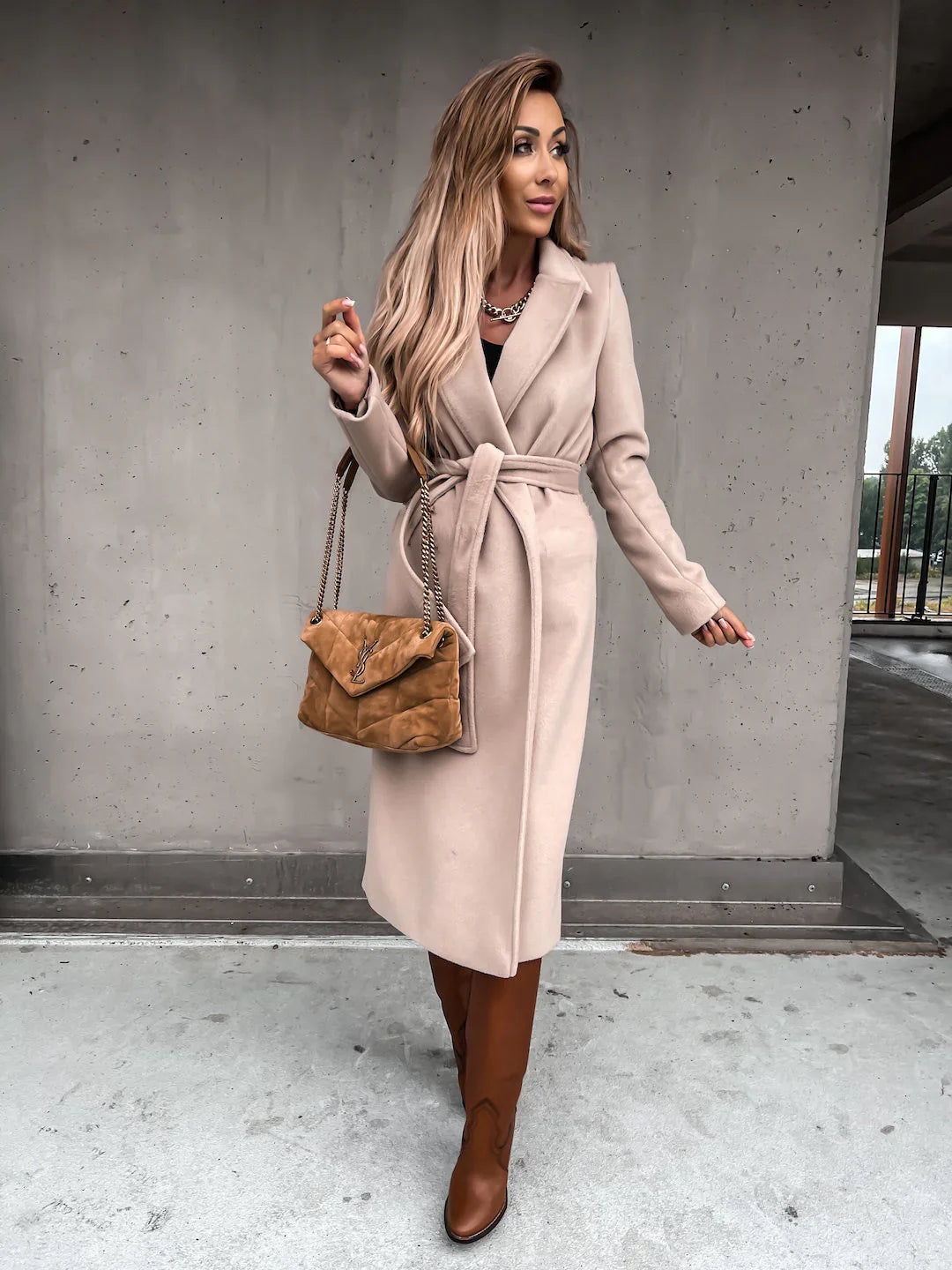 Woman in beige belted coat, brown knee-high boots, holding tan designer handbag against concrete wall